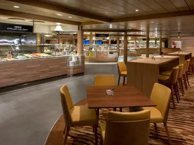 Geräumiges Buffetrestaurant der Spectrum of the Seas mit mehreren Essensstationen für kalte und warme Speisen sowie verschiedenen Sitzbereichen mit hellen Stühlen und Holztischen.