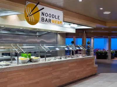Buffetbereich der Noodle Bar auf der Spectrum of the Seas. Eine Edelstahlküche und ein heller Speiseraum mit Meerblick durch Panoramafenster sind sichtbar. Speisen in Behältern stehen bereit.
