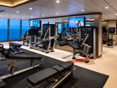 Modernes Fitnesscenter an Bord der Spectrum of the Seas mit Meerblick. Es bietet Kraft- und Cardiogeräte, Hantelbänke und Bildschirme. Große Fenster und Spiegel erzeugen ein offenes Raumgefühl.