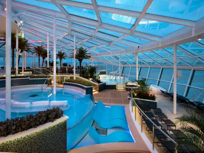 Überdachter Poolbereich, das Solarium, auf der Spectrum of the Seas. Mit blauem Wasser, Palmen, Liegestühlen, Glasdach und Fenstern zum Meer.