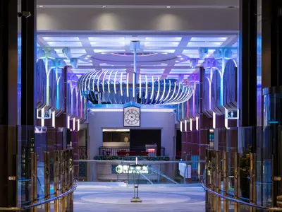 Die futuristische Innenhalle der Spectrum of the Seas. Eine blau leuchtende Deckeninstallation und farbige LED-Säulen prägen den Raum. Im Hintergrund eine klassische Uhr.