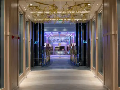 Gang auf der Spectrum of the Seas: Heller Boden, Holzwände, goldene Deckenverzierung. Farbig beleuchteter Bereich in der Ferne, Wegweiser.