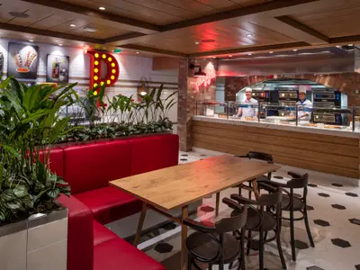Pizzeria an Bord der Spectrum of the Seas. Rote Sitzbänke, Holztische und Pflanzen im Essbereich. Hinten stehen zwei Köche an der Theke vor Pizzaöfen, ein rotes P-Schild leuchtet.