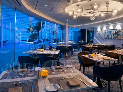Ein elegantes Restaurant auf der Spectrum of the Seas. Mehrere Tische sind gedeckt, dahinter eine Fensterfront zu einem blau illuminierten Poolbereich mit Palmen. Ein großer Kronleuchter hängt von der Decke, und ein abstraktes Gemälde ziert die Rückwand.
