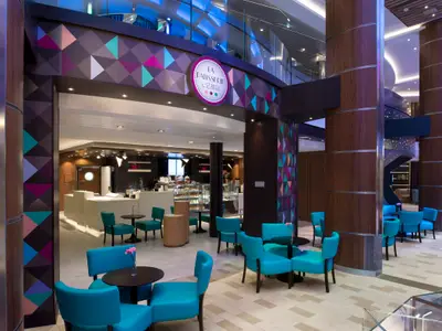 Interieur der La Patisserie auf der Spectrum of the Seas: tealblaue Stühle, Gebäcktheke und bunte, geometrische Wandmuster. Ein modernes Café an Bord.