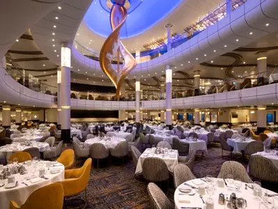 Der elegante, mehrstöckige Hauptspeisesaal der Spectrum of the Seas ist mit zahlreichen, gedeckten Tischen und einem großen, goldenen Kunstwerk im Zentrum zu sehen.