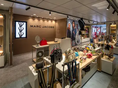 Blick in einen hellen Luxus-Boutique-Bereich an Bord der Spectrum of the Seas. Zu sehen sind Displayregale mit Handtaschen von Marc Jacobs und Michael Kors sowie Schuhen und Accessoires.