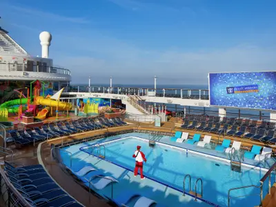 Blick auf das Hauptdeck der Spectrum of the Seas mit blauem Pool, bunten Wasserrutschen und einer großen Videoleinwand unter blauem Himmel. Eine Person steht im Pool.