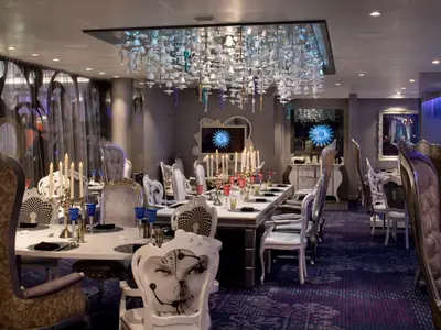 Restaurant auf der Spectrum of the Seas: surrealer Raum mit diversen Stühlen, gedeckten Tischen, Kerzen und Glocken-Schlüssel-Kronleuchter. Bildschirme zeigen blaue Muster.