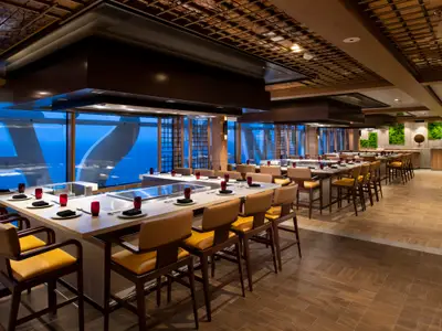 Das Teppanyaki-Restaurant an Bord der Spectrum of the Seas mit Tischen, Kochfeldern und Meerblick. Rote Gläser und gedeckte Plätze. Holzdecken und moderne Einrichtung.