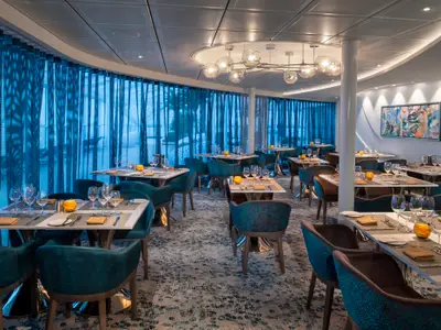 Innenansicht eines stilvollen Restaurants auf der Spectrum of the Seas. Mehrere Tische mit Stühlen, blaue Vorhänge an der Fensterfront, eine große Pendelleuchte und ein abstraktes Gemälde prägen den Raum.