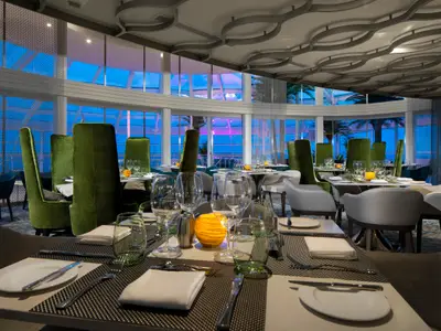 Ein elegantes Restaurant an Bord der Spectrum of the Seas. Mehrere Tische sind festlich gedeckt, umgeben von grünen Samtstühlen und großen Fenstern mit Blick nach draußen.