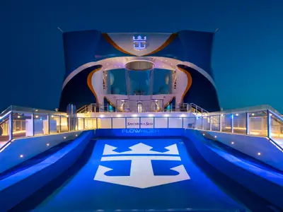 Oberdeck der Spectrum of the Seas bei Nacht. Beleuchteter FlowRider Surfsimulator im Vordergrund. Dahinter die gläserne Röhre des RipCord by iFly Skydiving-Simulators.