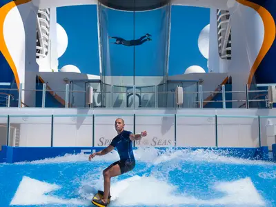 Funsport an Bord der Spectrum of the Seas: Ein Mann surft auf einer künstlichen Welle, darüber schwebt eine Person in einem Glas-Windtunnel. Blauer Himmel.
