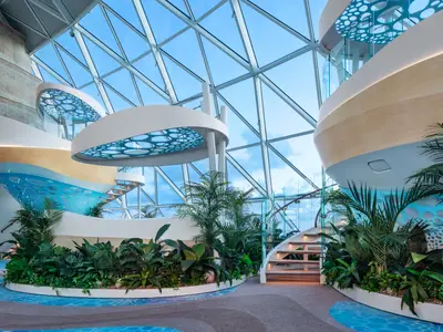 Innenbereich der Star of the Seas mit weitem Glasdach, üppigen Pflanzen, geschwungenen weißen Balkonen mit blauen Dekoren und eleganten Treppen.