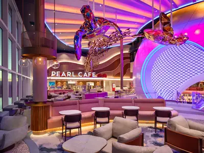 Innenansicht des Pearl Cafe auf der Star of the Seas. Der Raum ist hell und farbenfroh, mit rosa und violettem Licht. Große, bunte Schildkrötenskulpturen schweben über modernen Sitzgelegenheiten.