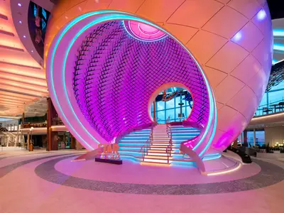 Innenansicht der Star of the Seas. Zentral: Die 'Pearl'-Skulptur mit farbiger Treppe und violettem Inneren. Oben sind Aktivitäten und Meerblick sichtbar. Der Schiffsname 'Star of the Seas' steht am Boden.