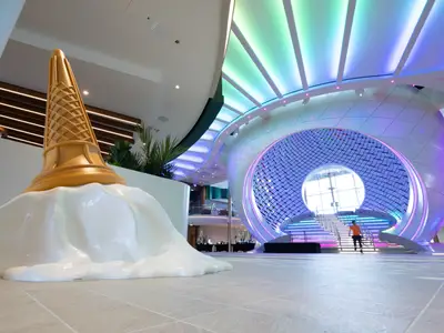 Modernes Interieur der Star of the Seas. Links eine riesige, umgefallene Eistüten-Skulptur. Rechts eine futuristische Halle mit farbig beleuchteter Treppe, die von einer Person betreten wird.