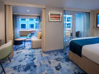 Helle Suite der Star of the Seas mit Bett, Sofa, Schreibtisch. Balkon blickt auf Schiff-Innenbereich mit anderen Kabinen. Blauer Teppich mit Muster.
