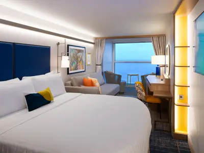 Moderne Kabine der Star of the Seas mit großem Bett, Sofa, Schreibtisch und weitem Meerblick. Die Einrichtung ist hell und farbenfroh in Blau, Weiß, Gelb und Orange.