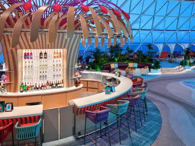 Eine geschwungene Bar mit bunten Hockern an Bord der Star of the Seas, umgeben von einem tropischen Ambiente. Der helle Bereich unter einem Glasdach zeigt Pflanzen, Lounge-Möbel und Getränke auf Regalen.