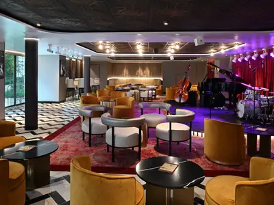 Innenansicht einer eleganten Jazz-Lounge mit einer Bühne auf der rechten Seite, ausgestattet mit Flügel, Schlagzeug und Kontrabass. Verschiedene Sitzbereiche mit bequemen Sesseln und Tischen füllen den Raum. Die Beleuchtung ist gedämpft und warm, an der Wand hängt ein Schwarz-Weiß-Foto eines Jazzmusikers.
