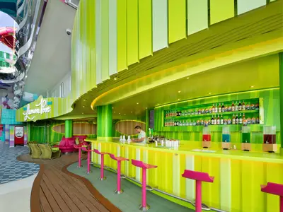 Lebhafte gelb-grüne Lemon Post Bar auf der Star of the Seas mit pinken Hockern, Barkeeper, Getränkeflaschen und bunten Spendergetränken.