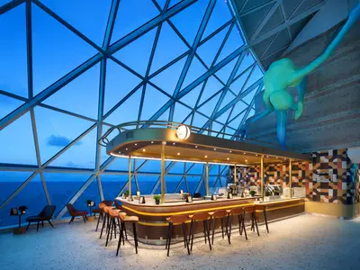 Interior einer modernen Bar auf der Star of the Seas. Große Fenster bieten Meerblick. Eine goldene Theke, Barhocker, Sitzgruppen und eine abstrakte blau-grüne Skulptur schmücken den Raum.