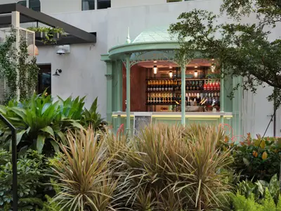 Eine mintgrüne Outdoor-Bar mit der Aufschrift „Bubbles“, gefüllt mit Champagnerflaschen. Davor üppige, grüne Pflanzen und ein Kreideschild. Im Hintergrund helle Gebäudewände.