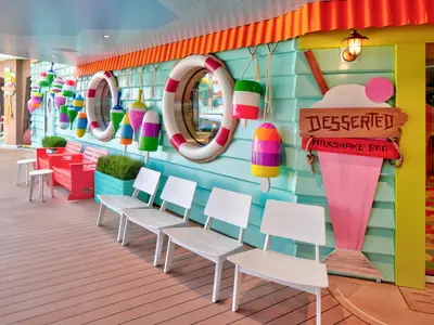 Der bunte Außenbereich eines Decks auf der Star of the Seas zeigt eine Milkshake Bar im maritimen Stil. Zu sehen sind bunte Bojen, Rettungsringe, hellblaue Holzverkleidung, rote Bänke und weiße Stühle.