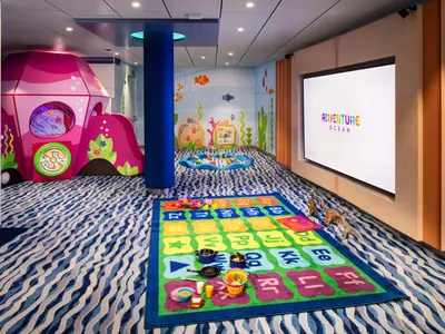 Blick in einen farbenfrohen Kinderclub an Bord der Star of the Seas mit einer großen pinken Spielburg, einem Alphabet-Teppich mit Spielzeug und Unterwasser-Wandmalereien. Ein großer Bildschirm zeigt 'ADVENTURE OCEAN'.