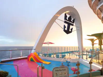 Star of the Seas: Bunter Kinder-Wasserpark mit Regenbogenrutsche und Wasserspielzeugen auf dem Deck. Ein großer Bogen mit Royal Caribbean Logo und das Meer im Sonnenuntergang sind zu sehen.