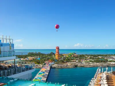 Die Star of the Seas liegt an einer tropischen Insel mit einem Wasserpark und einem roten Heißluftballon. Vom Deck aus sieht man den bunten Pier und das blaue Meer.