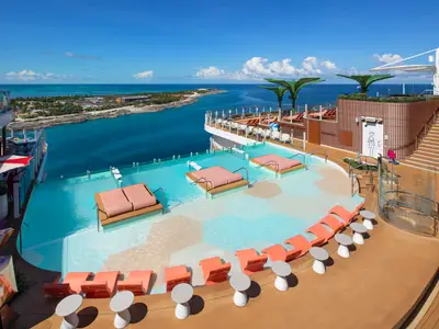 Sonniges Oberdeck der Star of the Seas: Pool mit Liegen und Stühlen. Blick auf das blaue Meer und eine Küste mit Gebäuden. Ein Bereich der Star of the Seas.