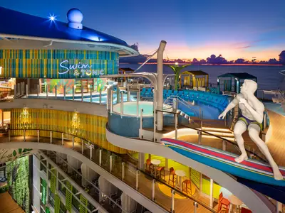 Das farbenfrohe Pooldeck der Star of the Seas bei Sonnenuntergang, mit der 'Swim & Tonic' Bar, mehreren Pools und einer Surfer-Statue. Das Meer ist im Hintergrund sichtbar.