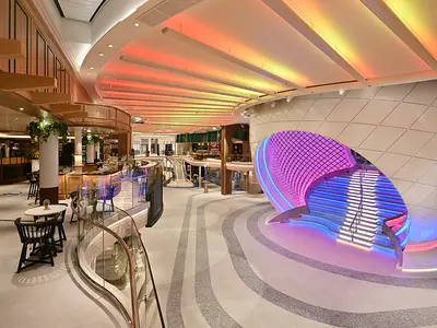 Innenansicht des modernen Atriums der Star of the Seas. Eine farbig leuchtende Skulptur mit Treppe ist rechts. Links ein Cafébereich mit Tischen und Glasbrüstung. Decke mit warmen Farblichtern.