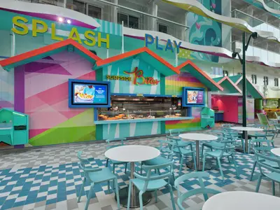 Ansicht des farbenfrohen Surfside Bites Gastronomiebereichs an Bord der Star of the Seas. Bunte Strandhütten-Theke, blaue Stühle, weiße Tische. Obere Decks erkennbar.
