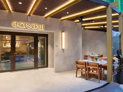 Heller Eingang des Restaurants Chops Grille mit leuchtendem Schriftzug. Davor moderne Tische und Stühle aus Holz für den Außenbereich. Der Innenraum ist durch Glas sichtbar.
