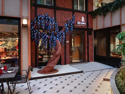 Eingang des japanischen Restaurants Izumi mit einer großen, kunstvollen Baumskulptur mit leuchtend blauen Blüten. Innen- und Außenbereiche mit Tischen sind zu sehen, der Boden ist hell gefliest.