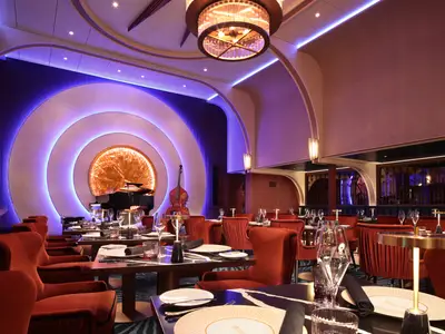 Elegantes Restaurant auf der Star of the Seas mit Bühne. Ein Flügel und Kontrabass stehen vor einer violett beleuchteten Wand. Rote Sessel und gedeckte Tische laden zum Verweilen ein.