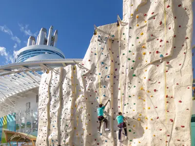 Zwei Personen klettern an der Außen-Kletterwand der Star of the Seas. Im Hintergrund des Royal Caribbean Schiffs sind dessen moderne Schornsteine und blauer Himmel zu erkennen.
