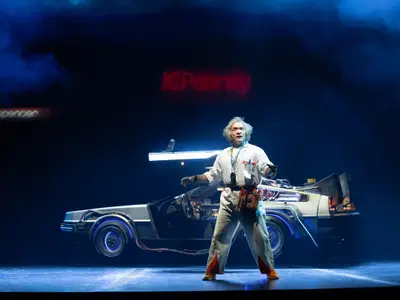 Ein Schauspieler als Doc Brown auf dunkler Bühne vor DeLorean-Auto. Leuchtende Logos "JCPenney", "spencer" und blauer Rauch im Hintergrund.