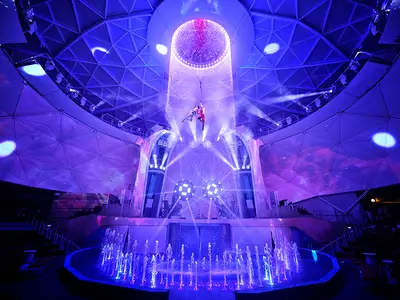 Blick in den beleuchteten AquaDome der Star of the Seas während einer Show. Zwei Artisten schweben unter einem Wasserfall, umgeben von leuchtenden Fontänen und Bühnenrequisiten.