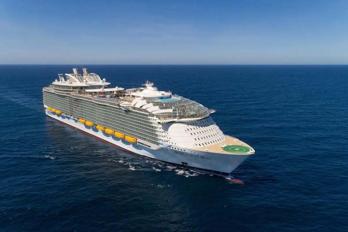 Eine Luftaufnahme zeigt die riesige Symphony of the Seas auf tiefblauem Meer unter wolkenlosem Himmel. Der Bug mit einem Helikopterlandeplatz und ihr Name sind deutlich sichtbar. Viele Rettungsboote sind an ihrer Seite erkennbar.