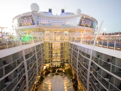 Blick in den Central Park der Symphony of the Seas. Man sieht begrünte Bereiche, beleuchtete Restaurants und unzählige Balkonkabinen, die das Atrium umgeben. Passagiere sind auf den oberen Decks erkennbar.