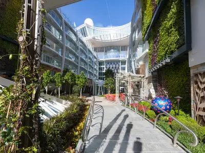 Der Central Park auf der Symphony of the Seas, mit üppigen vertikalen Gärten an den Gebäudewänden, Bäumen und einem Weg. Im Hintergrund moderne Architektur und eine blaue Kugel.