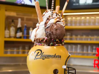 Ein riesiger Eisbecher mit Vanille- und Schokoladeneis, Sahne, Schokoladensauce, Waffelröllchen und bunten Streuseln. Im Hintergrund eine Bar mit Flaschen und Gläsern.