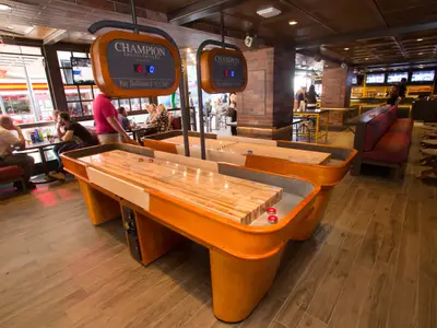 Zwei orangefarbene Shuffleboard-Tische mit Anzeigetafeln im Entertainment-Bereich der Symphony of the Seas. Gäste spielen oder entspannen an Tischen; eine Bar ist im Hintergrund.