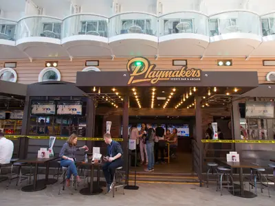 Detailansicht der Playmakers Sports Bar & Arcade auf der Symphony of the Seas mit Gästen an Tischen im Freien und im Innenbereich. Mehrere Bildschirme zeigen ein Basketballspiel. Im oberen Bereich sind Kabinenbalkone des Schiffs zu sehen.