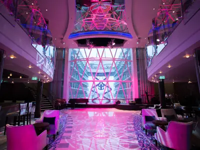 Blick ins stilvolle Atrium der Symphony of the Seas. Pinkes Licht beleuchtet Bühne, Flügel, Lounge-Sessel und Bar. Durch die Glasfront sind Außendecks und der Schiffsname sichtbar.